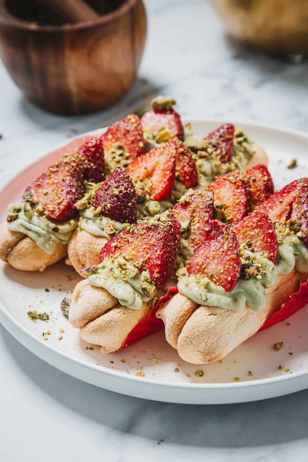 Eclairs topped with green cream, sliced strawberries, and crushed pistachios on a white plate&mdash;like a No-Bake Strawberry Pistachio Ladyfingers Dessert reinvented in &eacute;clair form.