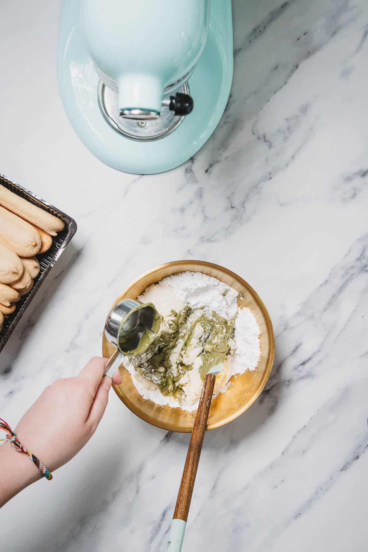 A hand pours liquid into a mixing bowl of flour beside a blue stand mixer on a marble counter, preparing ingredients for a No-Bake Strawberry Pistachio Ladyfingers Dessert.