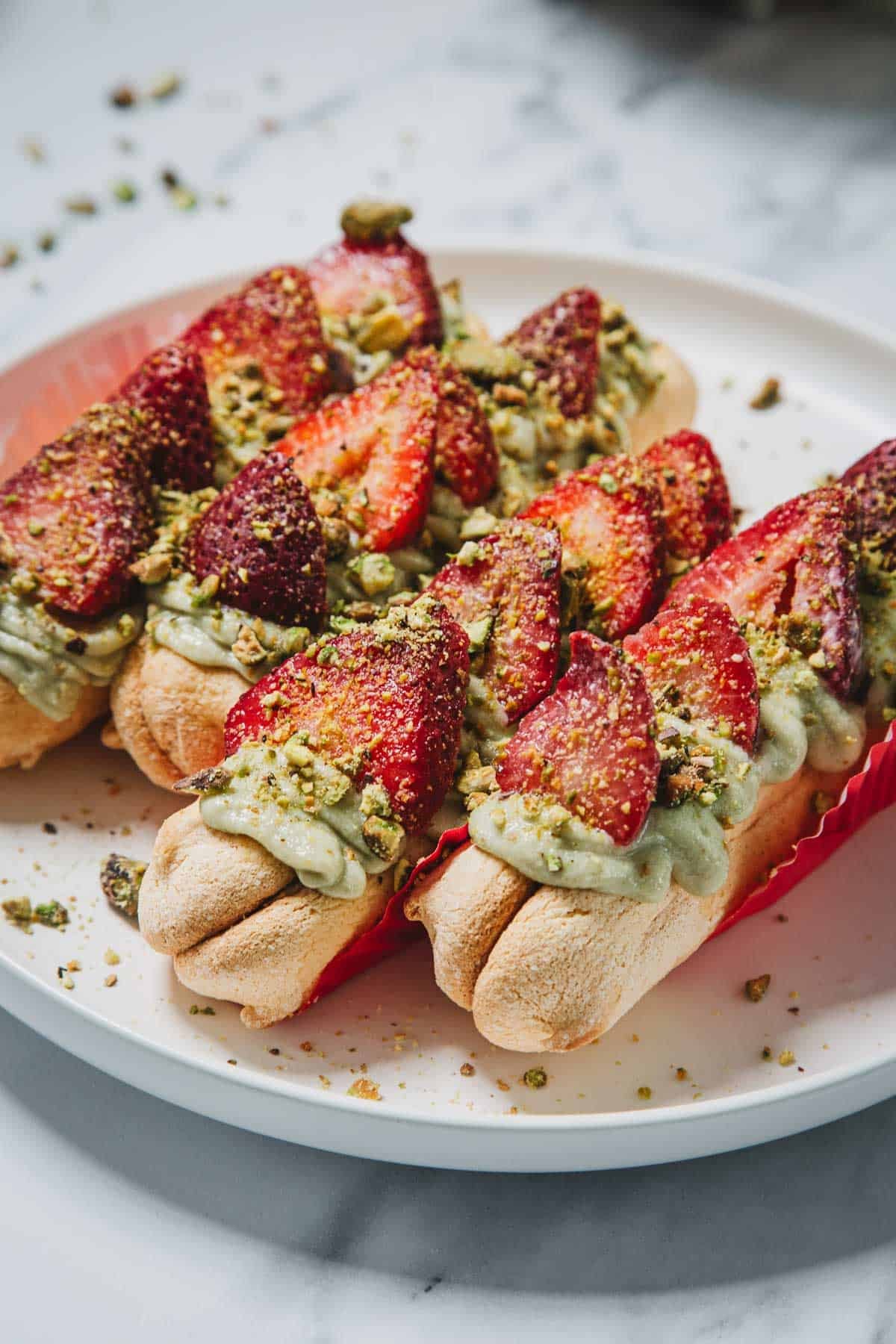 Three eclairs topped with green cream, sliced strawberries, and crushed pistachios evoke the flavors of a No-Bake Strawberry Pistachio Ladyfingers Dessert on a white plate.