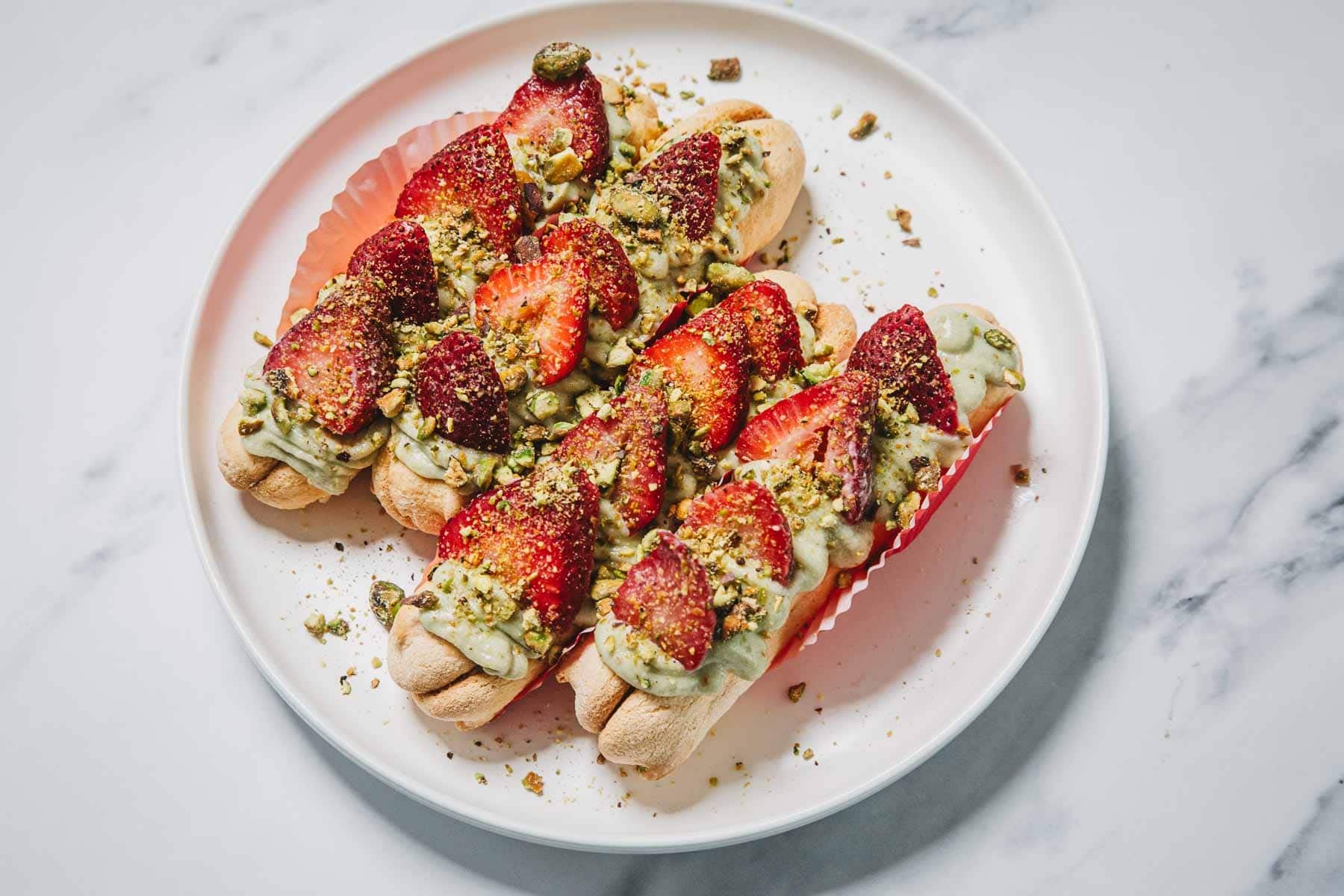 No-Bake Strawberry Pistachio Ladyfingers Dessert features ladyfingers topped with green cream, sliced strawberries, and crushed pistachios on a white plate.