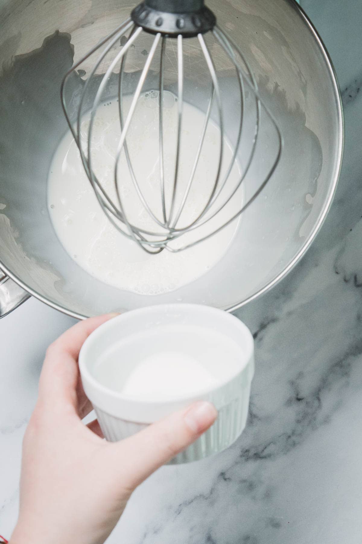 A hand holds a small white bowl of sugar over a mixer bowl with cream and a whisk attachment, preparing the base for a No-Bake Strawberry Pistachio Ladyfingers Dessert.