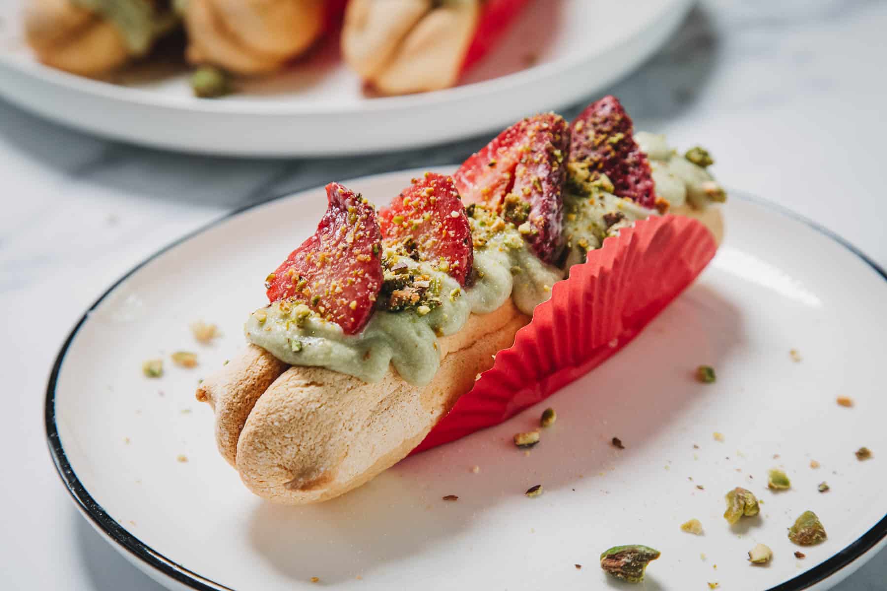 A No-Bake Strawberry Pistachio Ladyfingers Dessert in a red wrapper, topped with green cream, sliced strawberries, and crushed pistachios on a plate.
