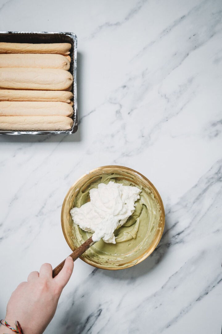A hand mixes cream into a green batter in a bowl beside a tray of ladyfinger cookies, preparing a No-Bake Strawberry Pistachio Ladyfingers Dessert on a marble surface.