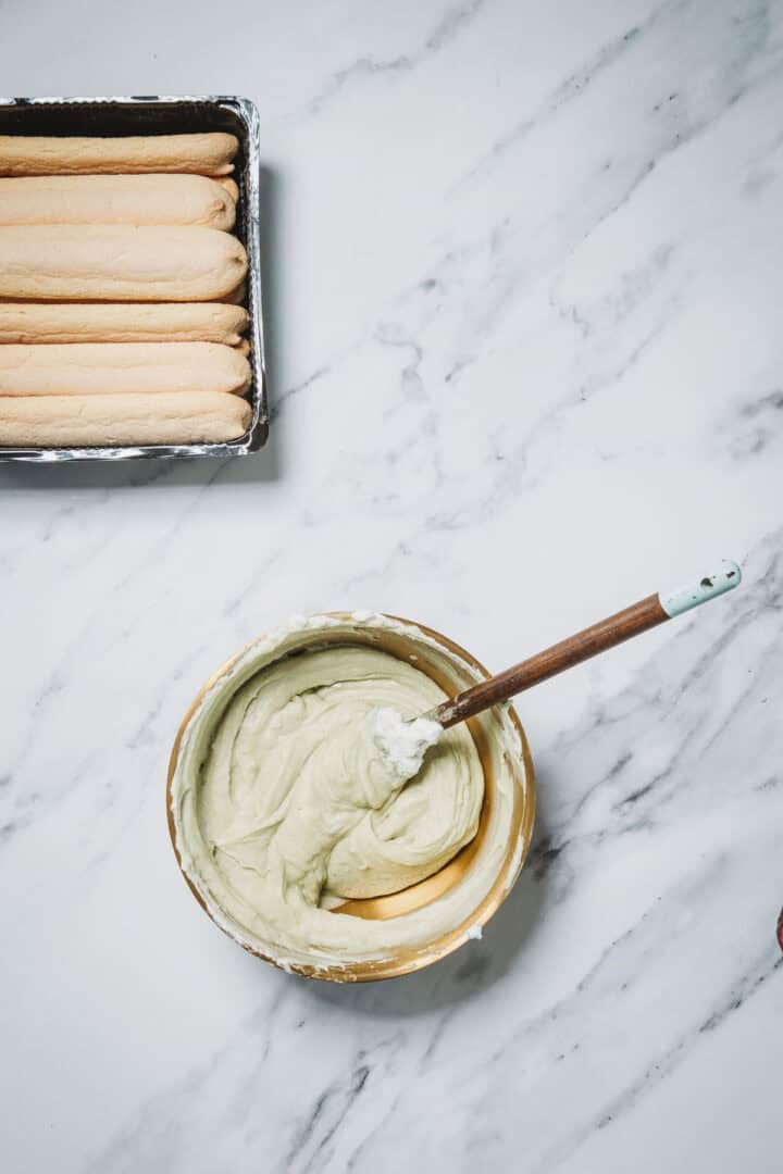 Bowl of creamy mixture with a spatula beside a tray of ladyfinger cookies on a marble surface, perfect for assembling a No-Bake Strawberry Pistachio Ladyfingers Dessert.