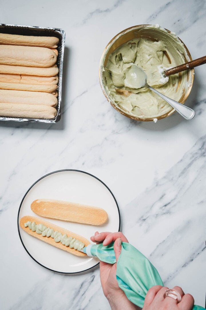 A hand pipes cream onto a ladyfinger for this No-Bake Strawberry Pistachio Ladyfingers Dessert, with more ladyfingers and a bowl of cream nearby on a marble surface.