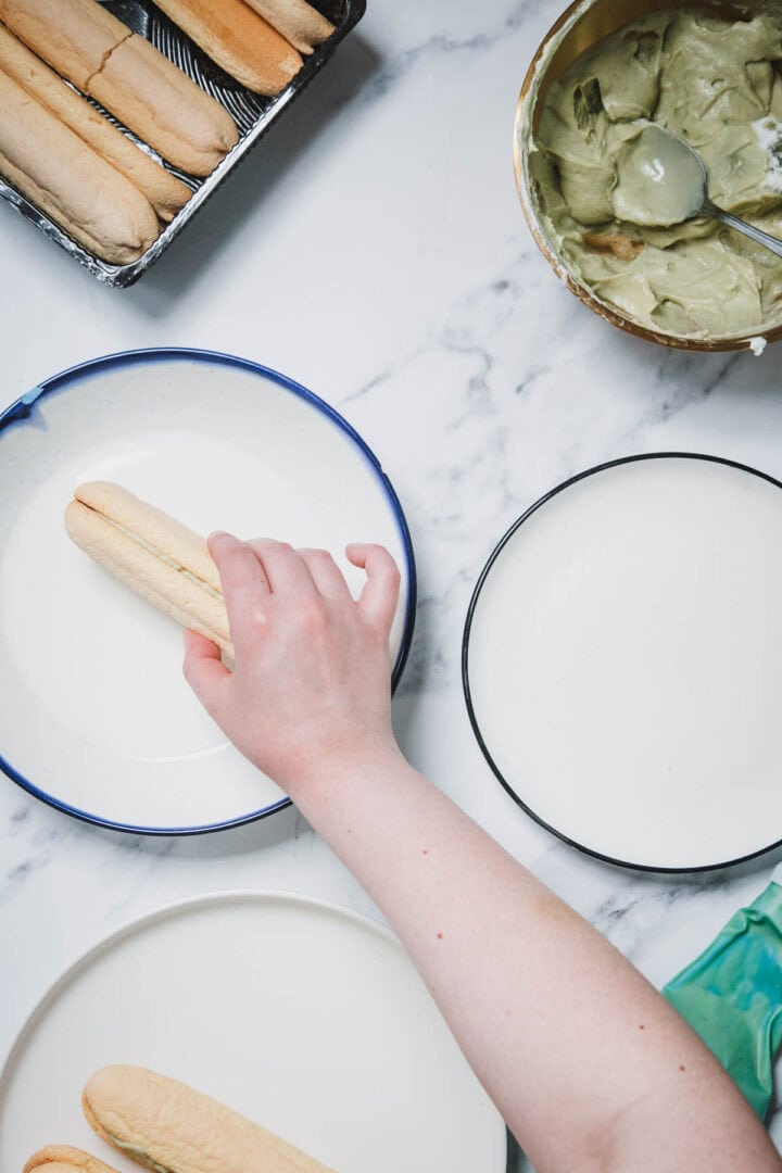 A hand dips a ladyfinger biscuit into a bowl of liquid beside plates and a bowl of cream on a marble surface, preparing the No-Bake Strawberry Pistachio Ladyfingers Dessert.