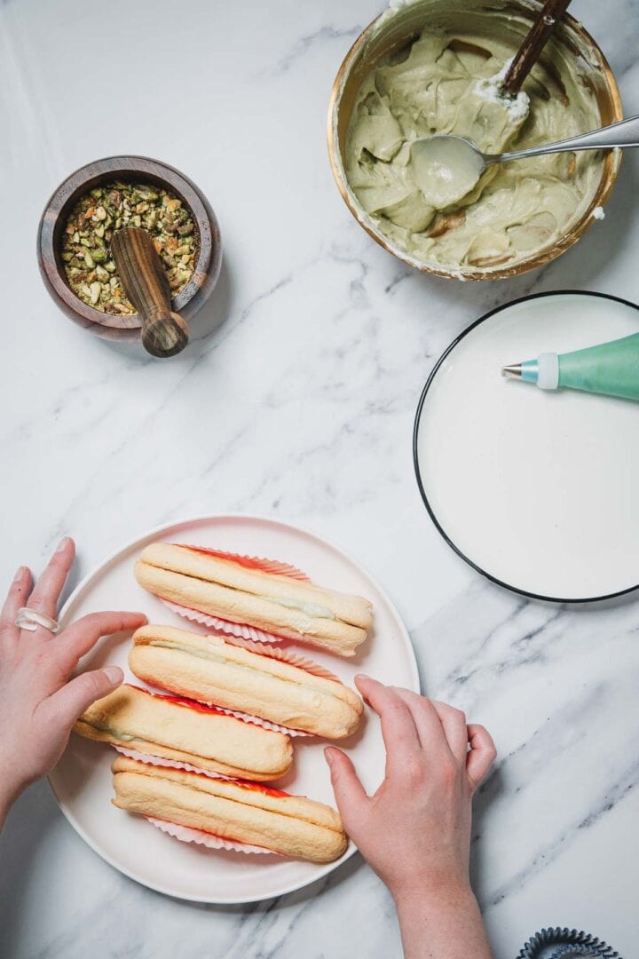 Hands arranging ladyfingers on a plate for a No-Bake Strawberry Pistachio Ladyfingers Dessert, with bowls of cream, chopped nuts, and a piping bag nearby on the marble table.
