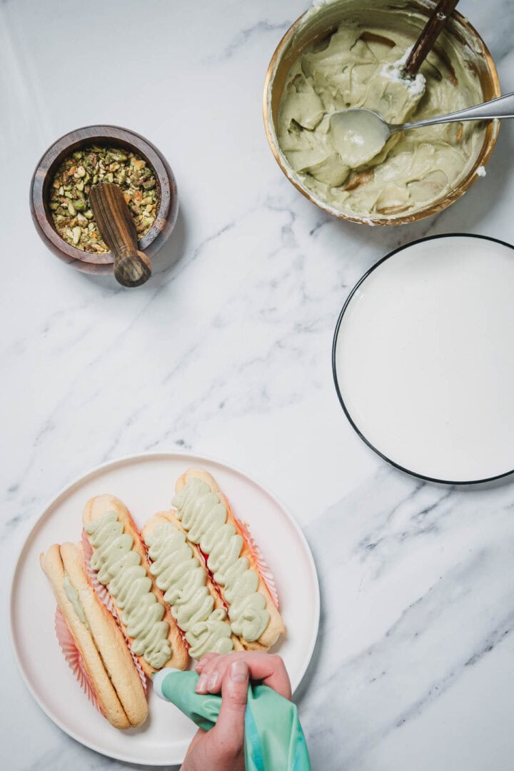 A hand pipes green cream onto three ladyfingers on a plate, preparing a No-Bake Strawberry Pistachio Ladyfingers Dessert, with bowls of topping and filling nearby.