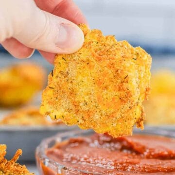 A hand dips a crispy, breaded zucchini chip into a bowl of marinara sauce.