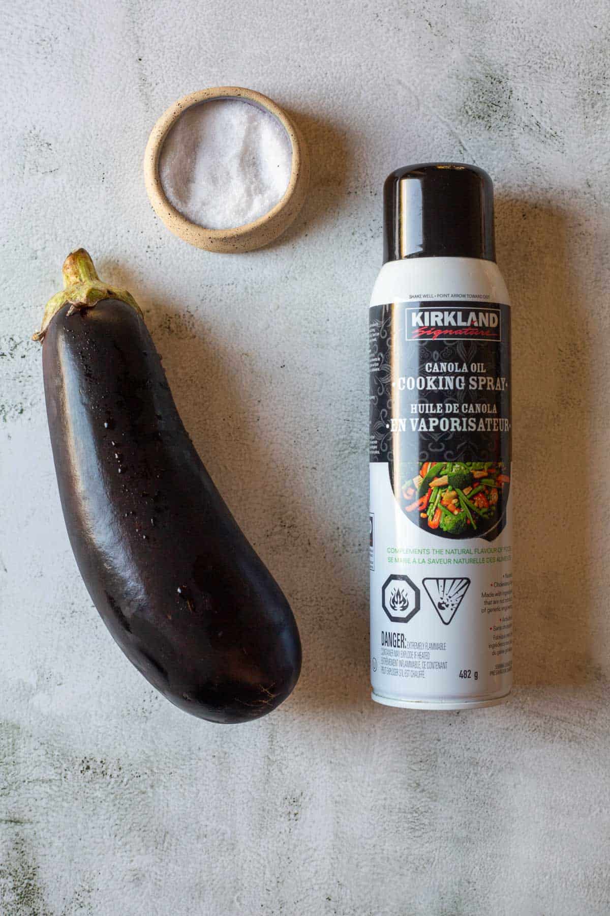 An eggplant, a small bowl of salt, and a can of Kirkland canola oil cooking spray on a gray surface—perfect for making crispy air fryer eggplant.