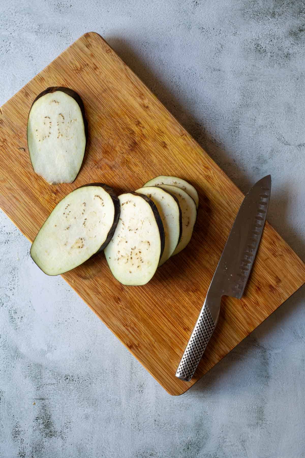 Sliced eggplant and a knife on a wooden cutting board, ready to become delicious air fryer eggplant, placed on a light gray surface.