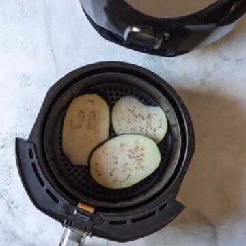 Three slices of air fryer eggplant sit in an air fryer basket next to the sleek black appliance on a light surface.