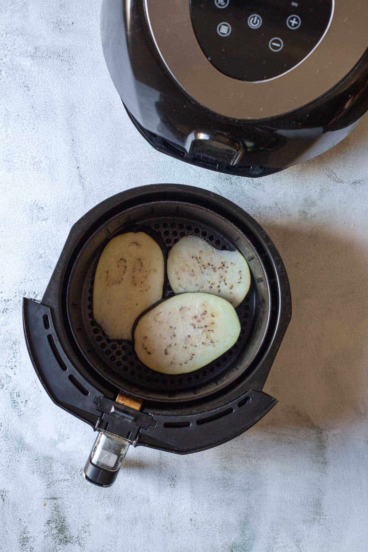Three slices of air fryer eggplant sit in an air fryer basket next to the sleek black appliance on a light surface.