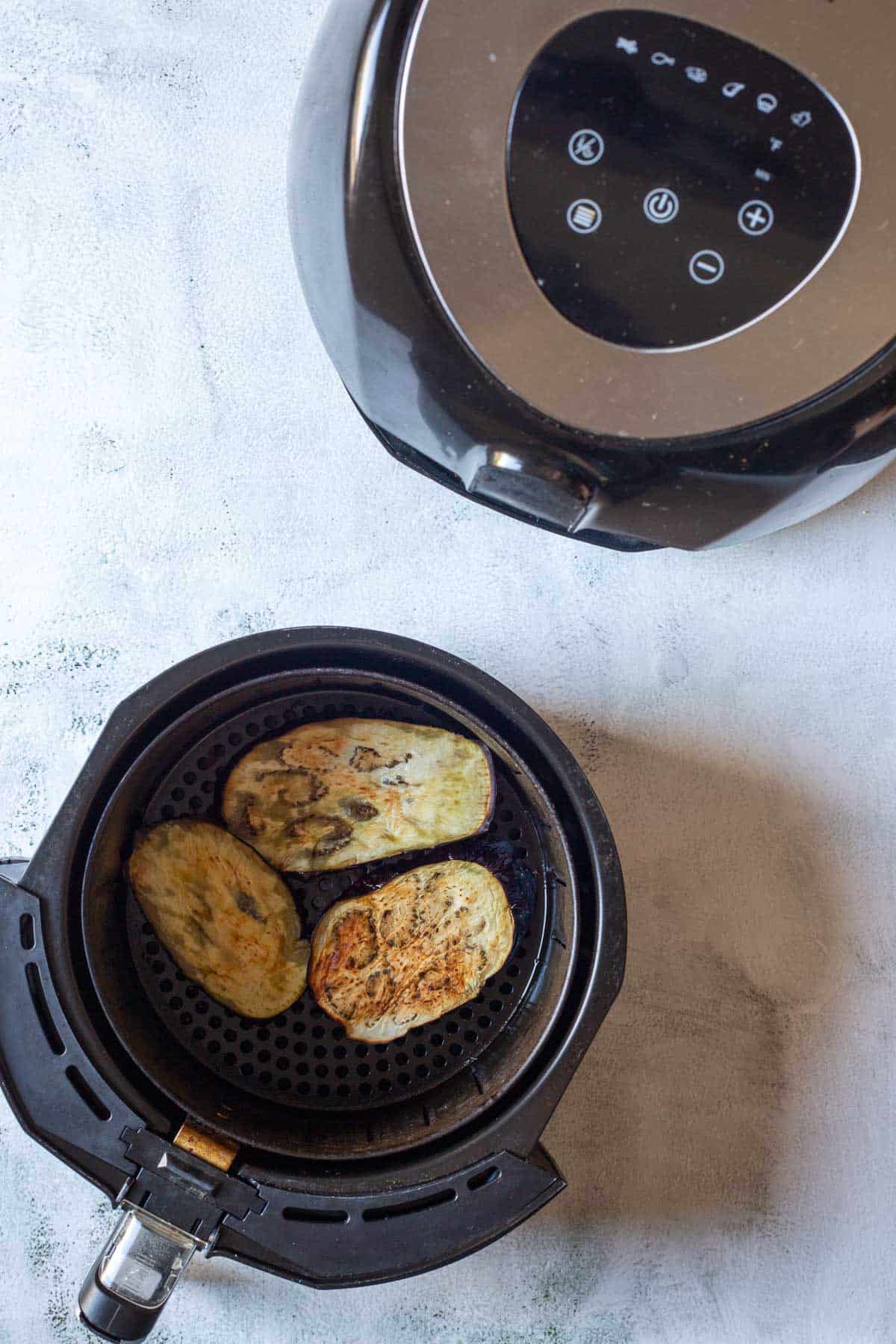 Slices of air fryer eggplant rest in the basket, perfectly cooked and ready to enjoy, on a light countertop.