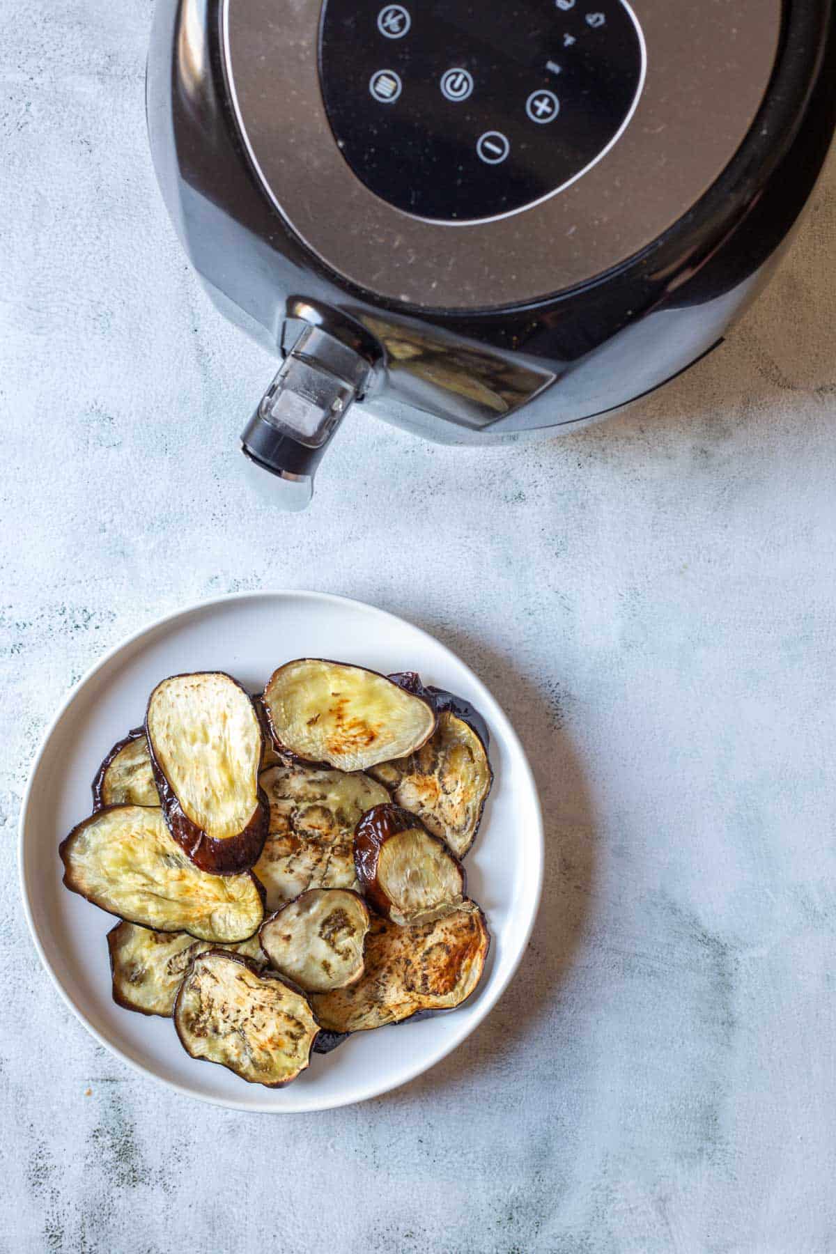 A bowl of crispy air fryer eggplant slices sits beside a black air fryer on a light gray surface.