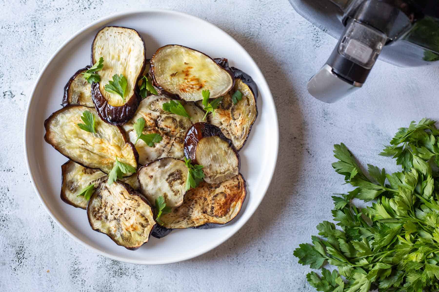 A plate of crispy air fryer eggplant slices garnished with fresh parsley sits beside an air fryer and a bunch of parsley.