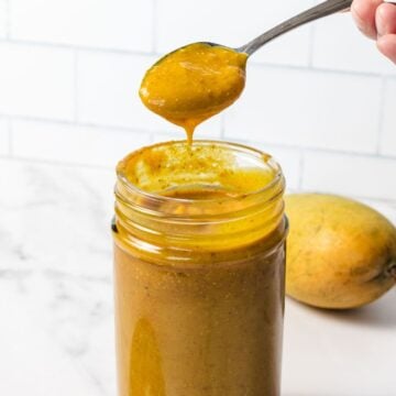 Spoon drizzling golden mango chutney into a glass jar, with a fresh mango in the background.