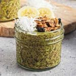 A jar of green chia pudding topped with banana slices, granola, shredded coconut, and dried berries.