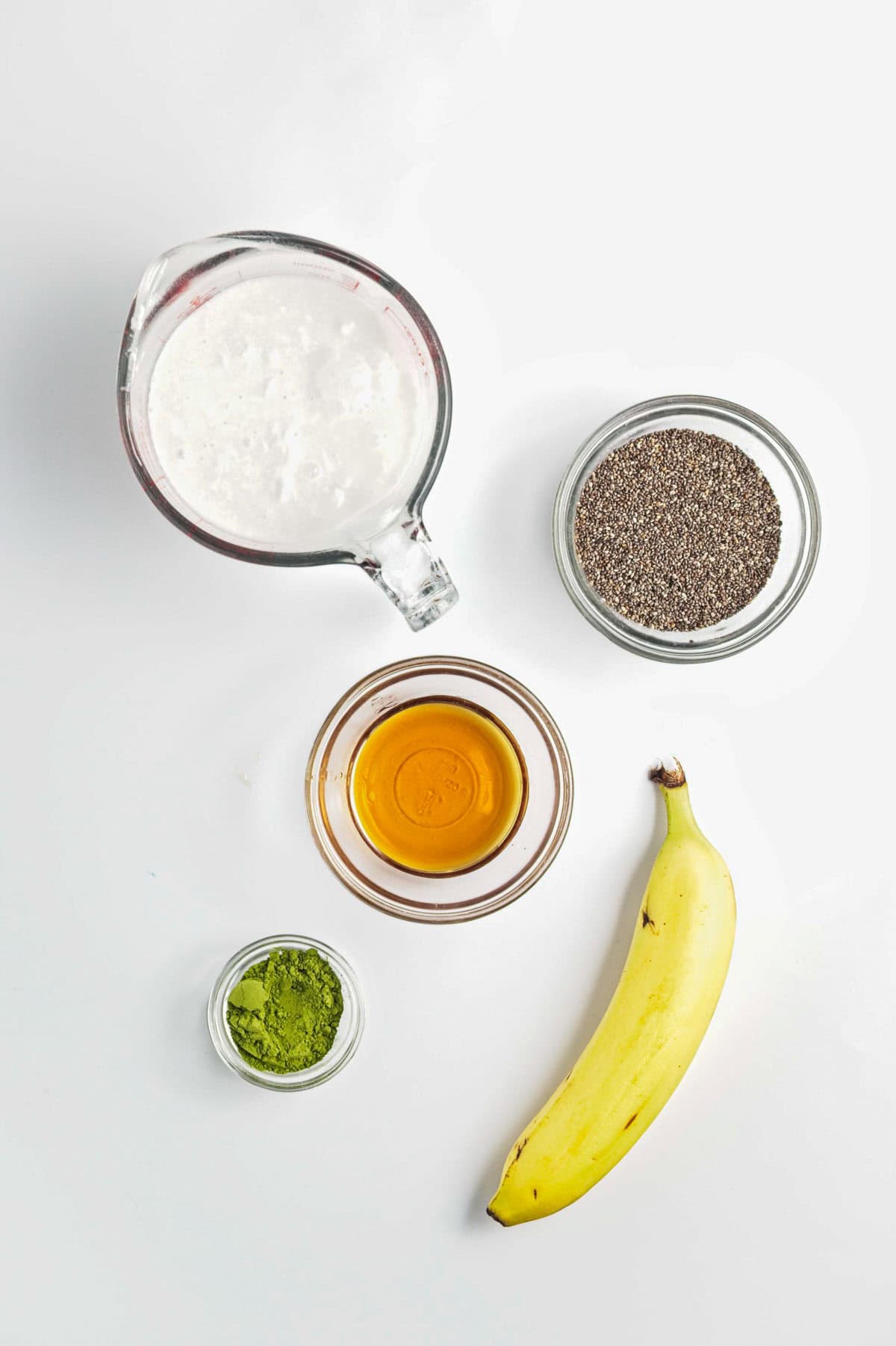 A banana, chia seeds, matcha powder, honey, and a jug of milk arranged on a white background&mdash;perfect ingredients for Banana Matcha Chia Pudding.