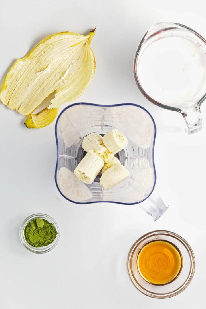 Sliced banana in a blender with a banana peel, matcha powder, milk, honey, and chia seeds on a white background&mdash;perfect for making Banana Matcha Chia Pudding.