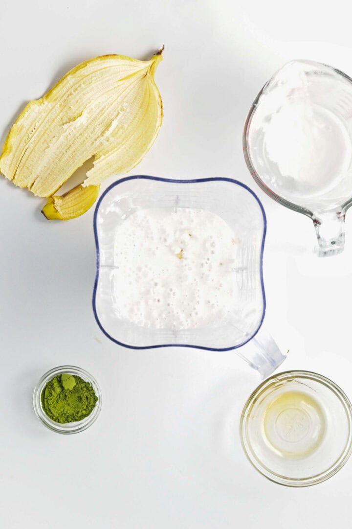Blender with smoothie, banana peel, matcha powder, measuring cup, and bowl on a white surface&mdash;perfect for making Banana Matcha Chia Pudding.