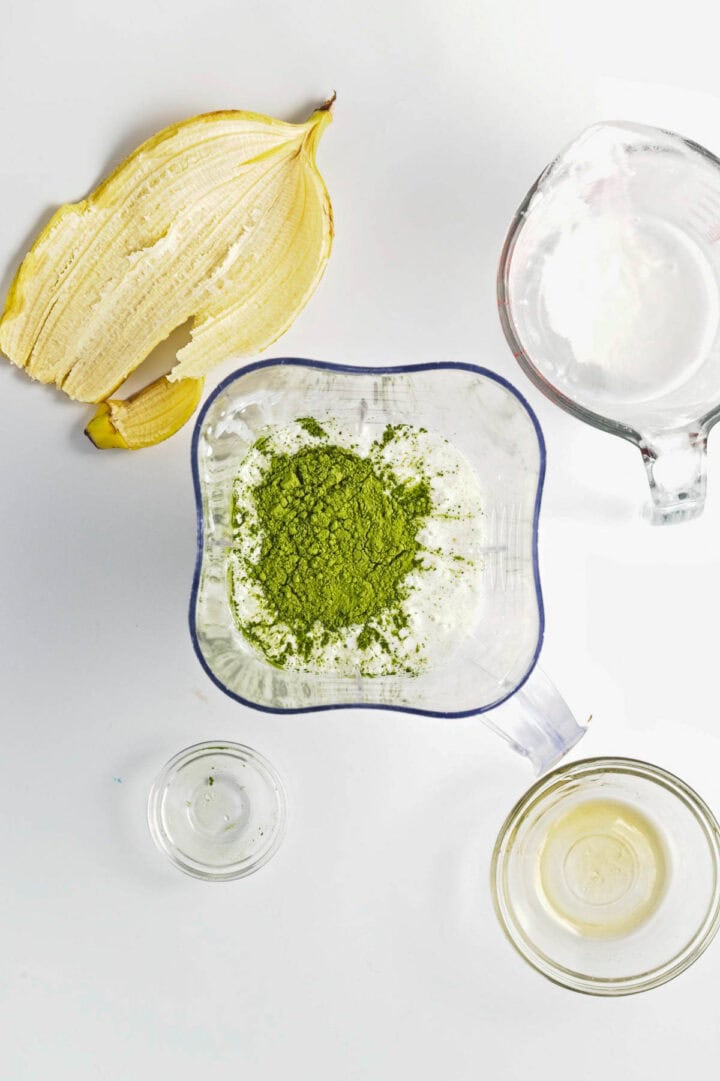 Matcha powder, banana peel, a blender, a glass of water, and a small bowl of liquid are arranged on a white surface&mdash;perfect ingredients for whipping up Banana Matcha Chia Pudding.