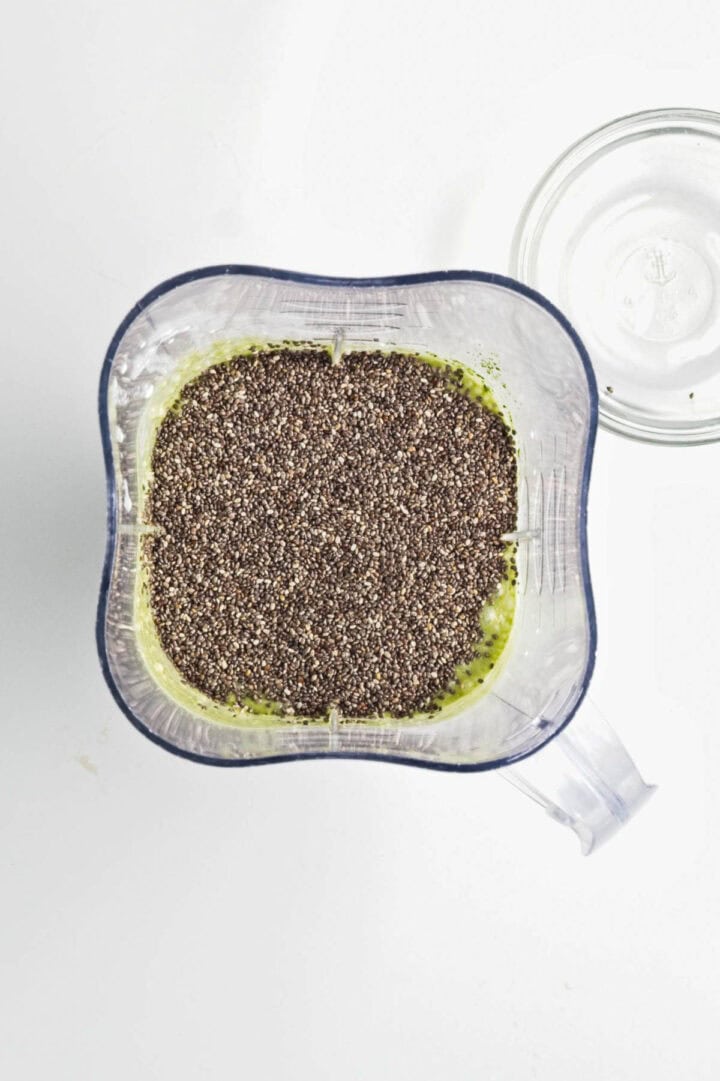 Top view of a blender with green smoothie and a layer of chia seeds on top, next to an empty glass&mdash;perfect for making Banana Matcha Chia Pudding.
