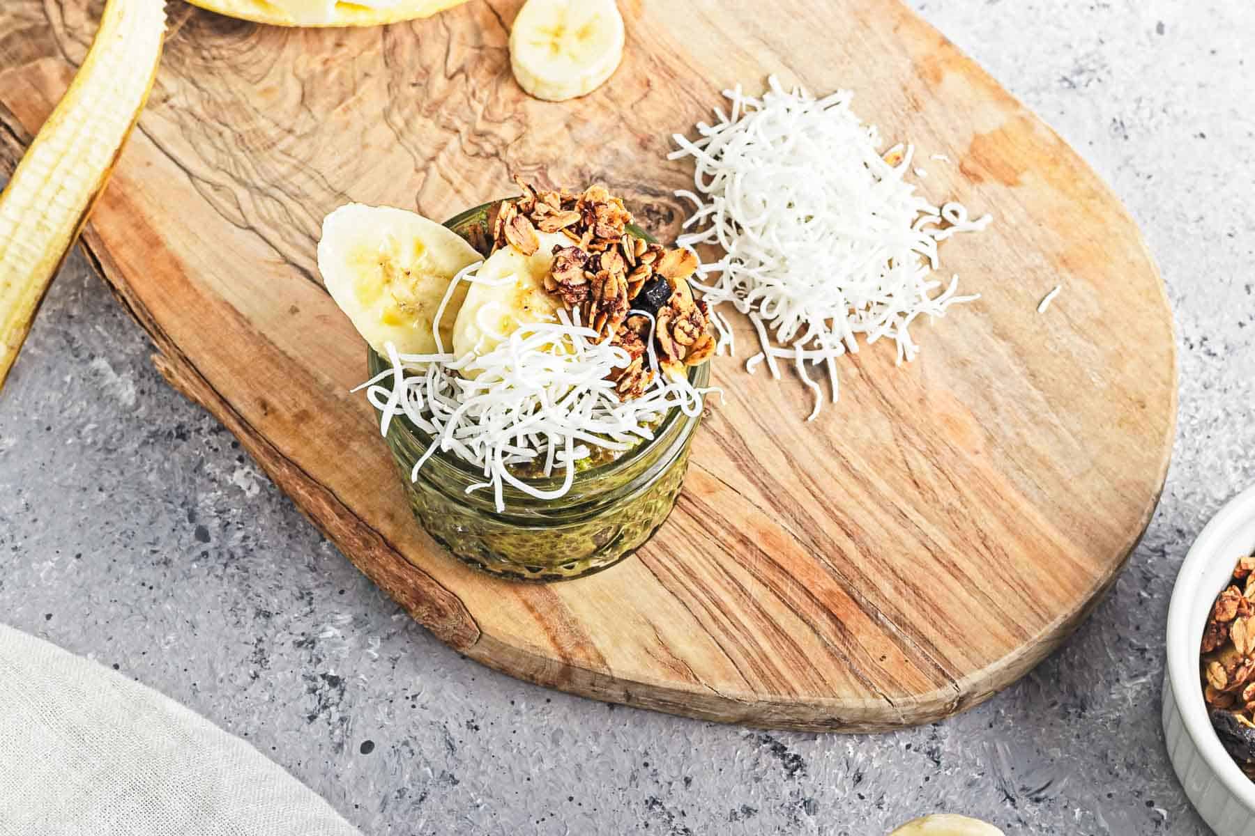 A glass jar of Banana Matcha Chia Pudding topped with granola, banana slices, and shredded coconut sits on a wooden board.