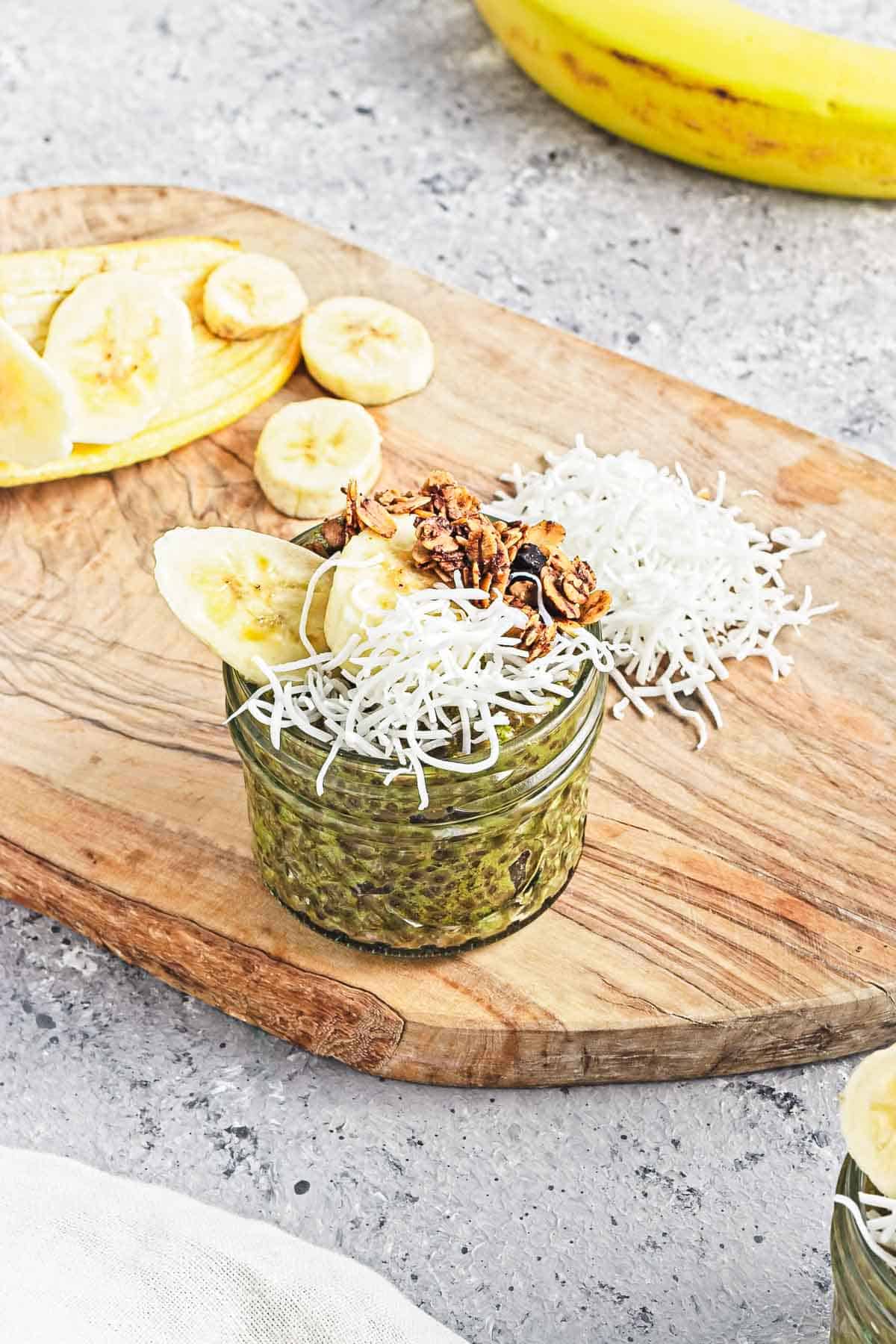 A glass jar of Banana Matcha Chia Pudding topped with shredded coconut, granola, and banana slices sits on a wooden board.