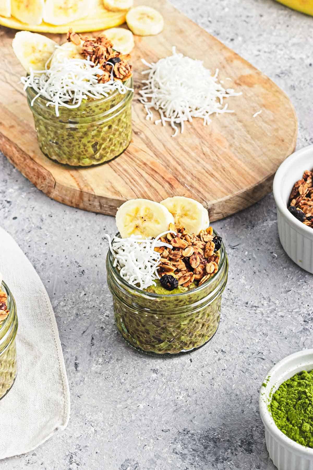A mason jar filled with Banana Matcha Chia Pudding, topped with fresh banana slices, granola, and shredded coconut.