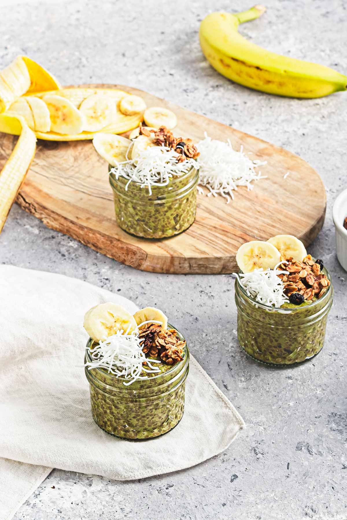 Three jars of Banana Matcha Chia Pudding topped with banana slices, shredded coconut, and granola, with fresh bananas in the background.