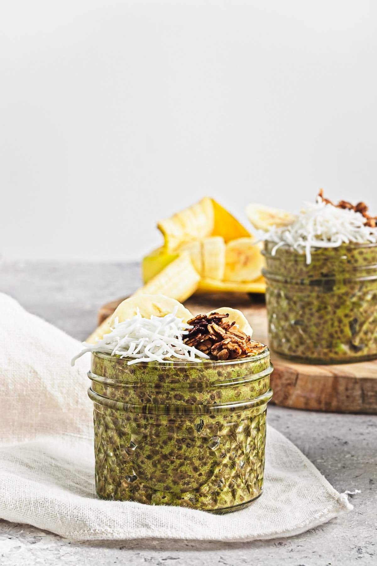 A jar of Banana Matcha Chia Pudding topped with shredded coconut, banana slices, and nuts, with fresh bananas in the background.