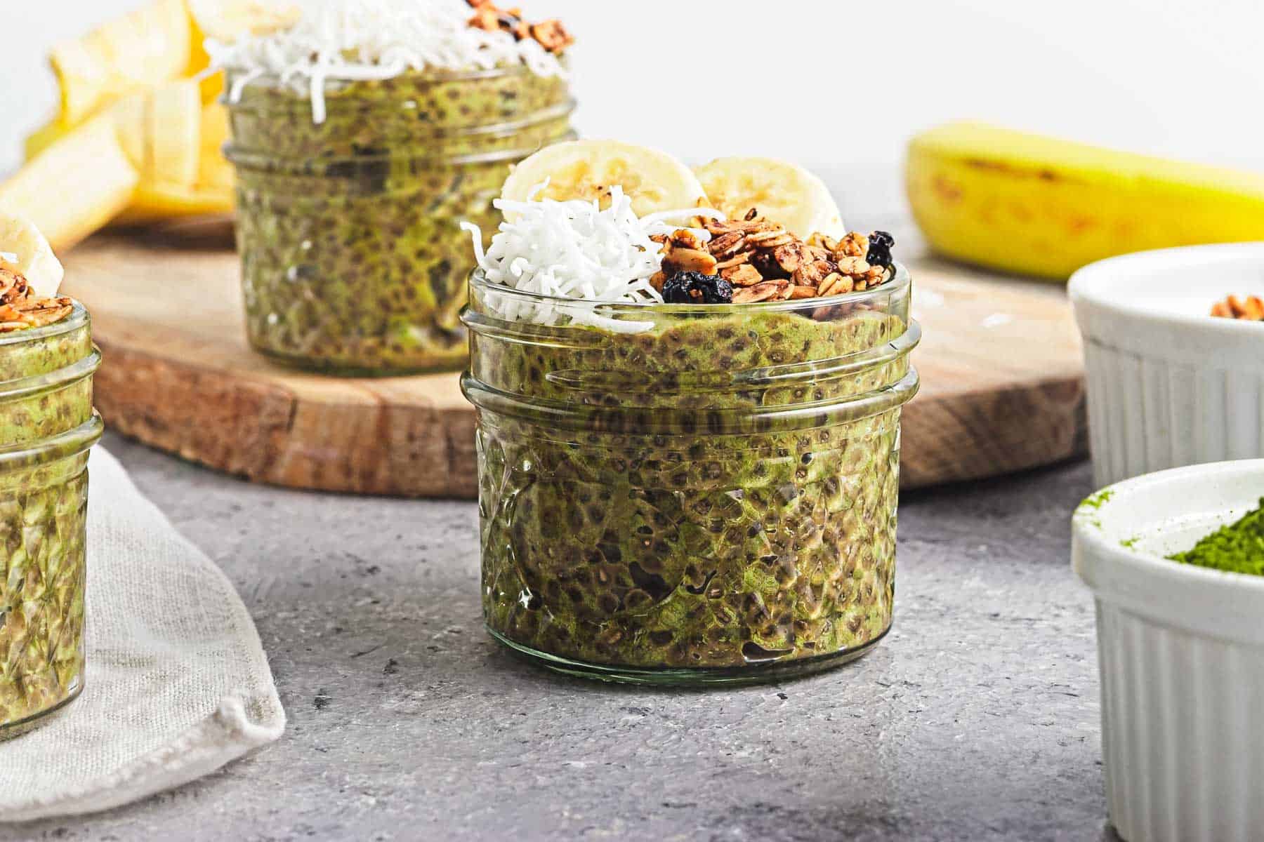 Jar of green chia pudding topped with banana slices, granola, and shredded coconut on a gray surface.