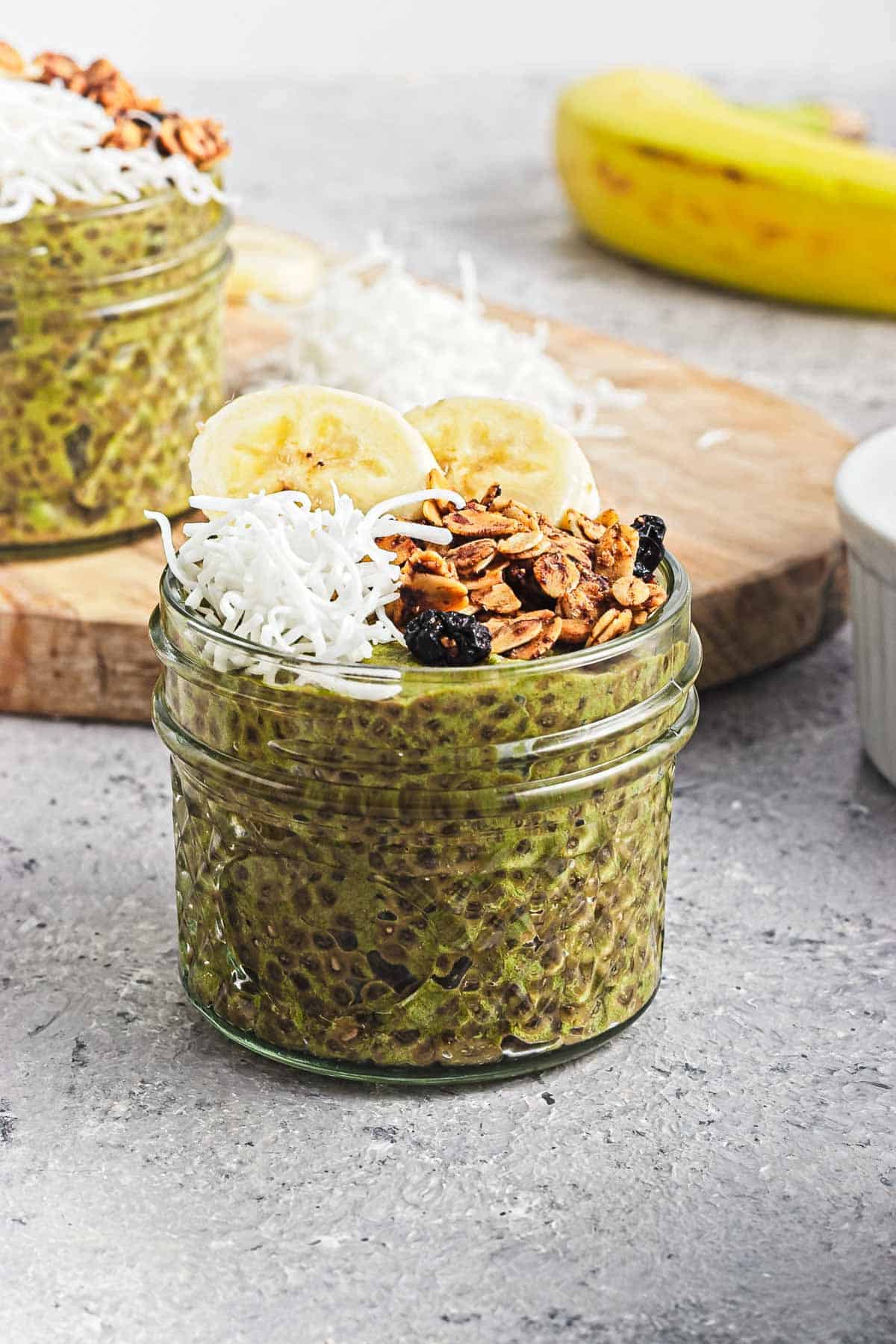 A jar of Banana Matcha Chia Pudding topped with banana slices, granola, and shredded coconut sits on a gray surface.