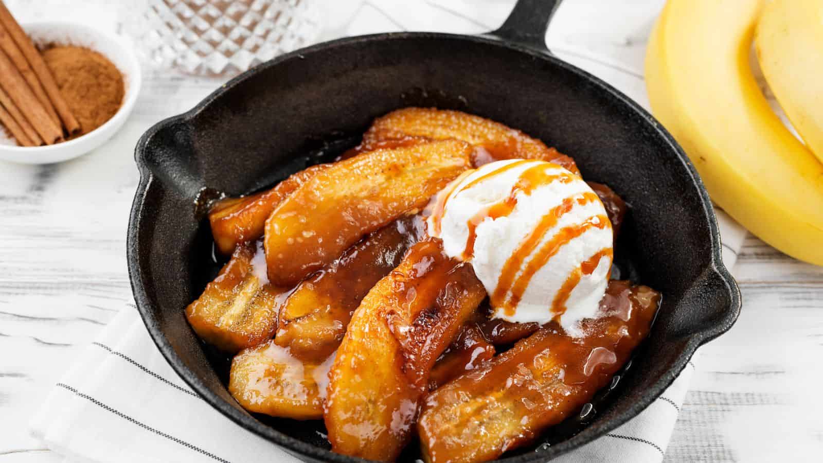 A cast iron skillet filled with caramelized banana slices, topped with a scoop of vanilla ice cream and drizzled with caramel sauce. A bowl of cinnamon and bananas are visible in the background.