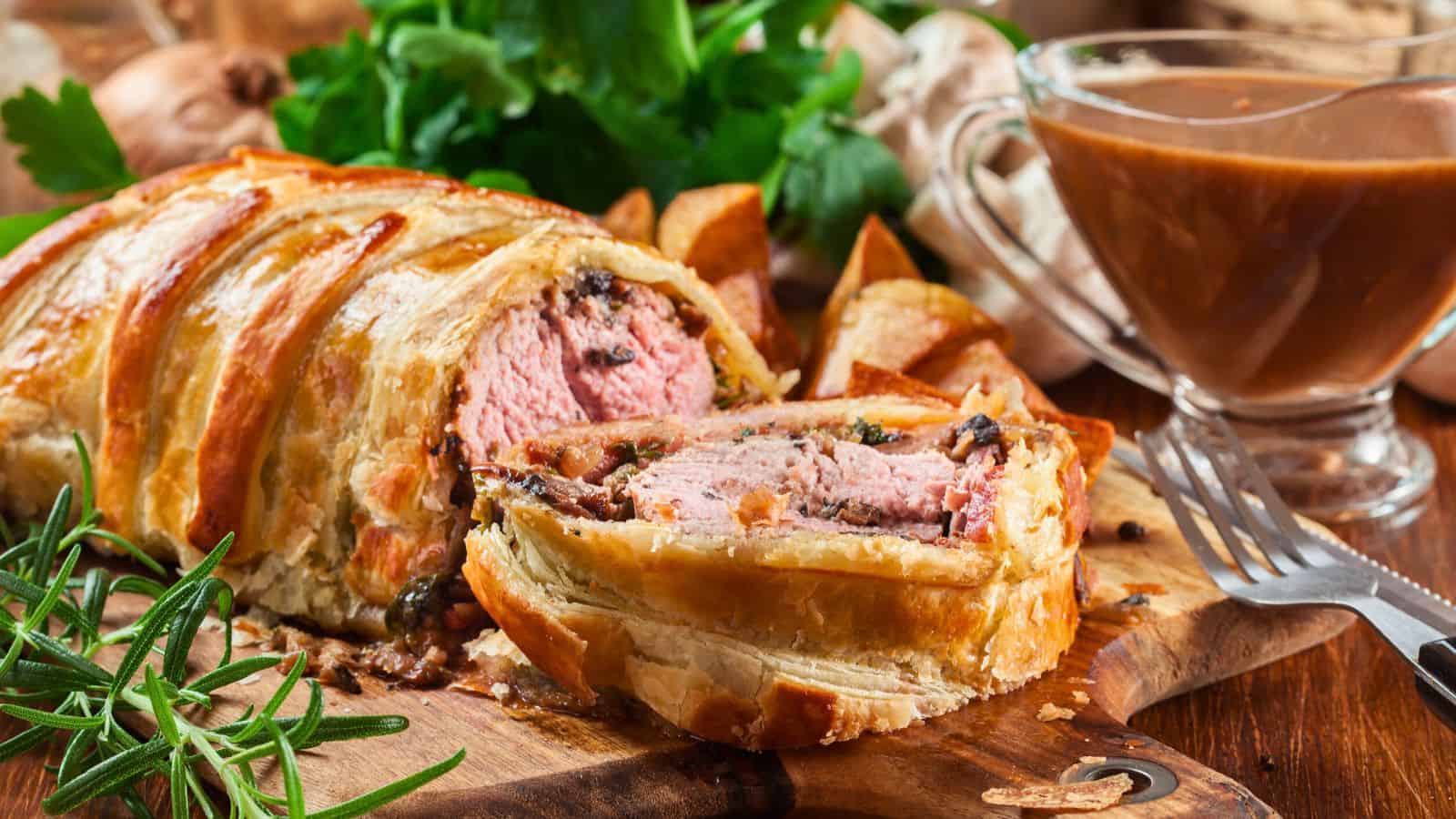 A sliced beef Wellington with a golden-brown pastry crust is displayed on a wooden board with herbs, roasted potatoes, and a glass gravy boat in the background.