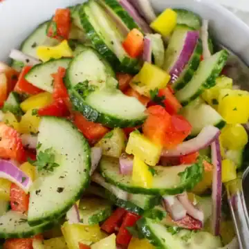 A bowl of fresh cucumber salad with red onion, bell peppers, and herbs, served with a spoon.