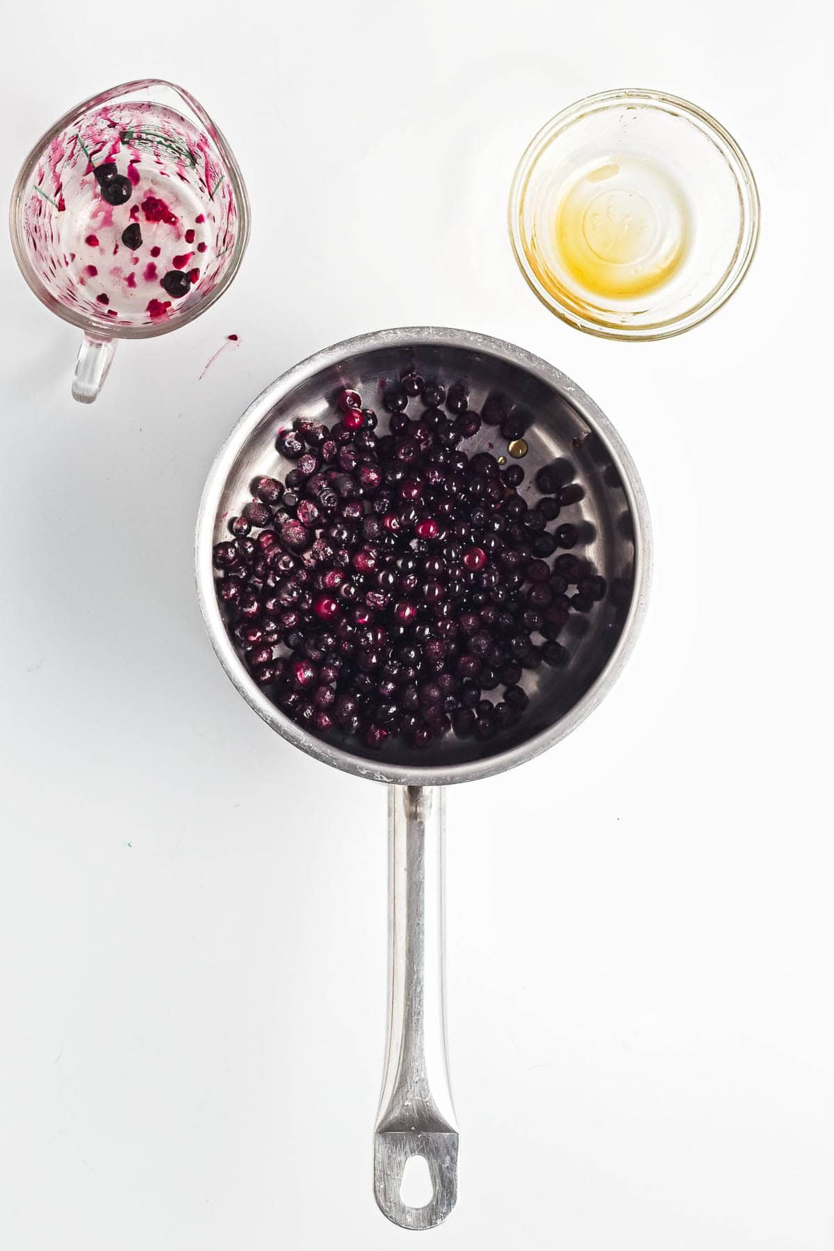 A saucepan with blueberries sits beside a glass with berry residue and a small bowl of liquid on a white surface, perfect for preparing Berry Chia Pudding.