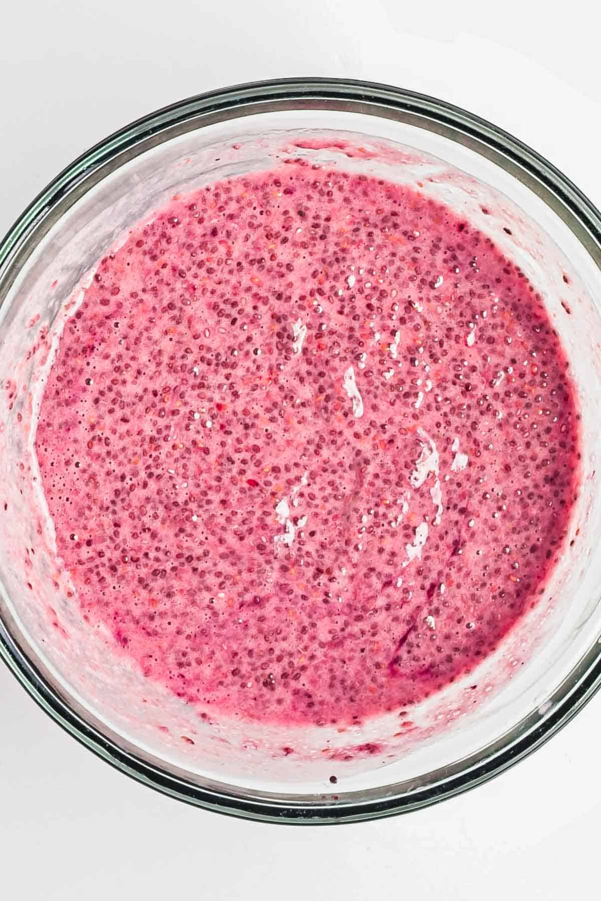 A glass bowl filled with bright pink Berry Chia Pudding mixture sits on a white surface.