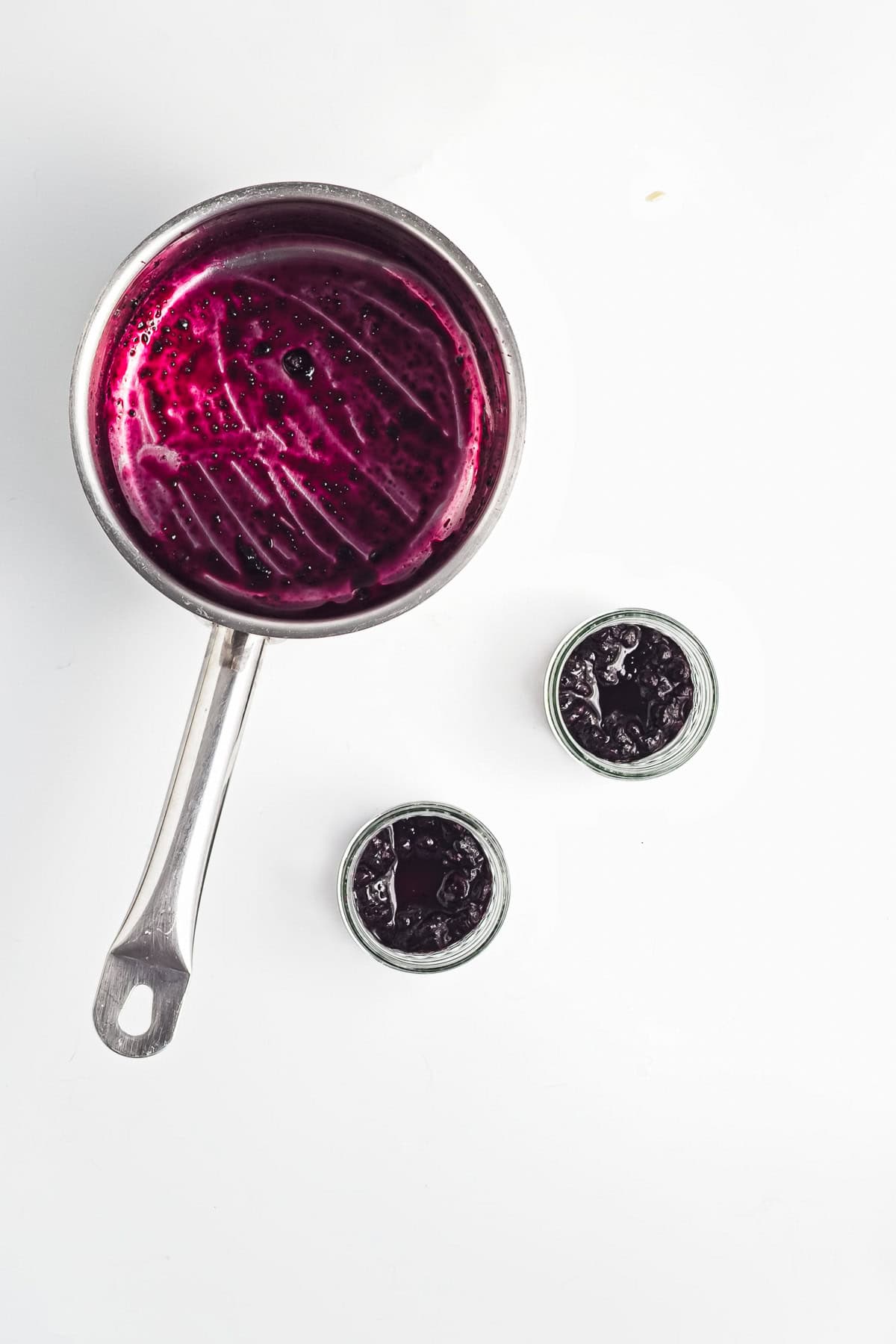 A saucepan with purple jam and two small jars of Berry Chia Pudding sit on a white surface.
