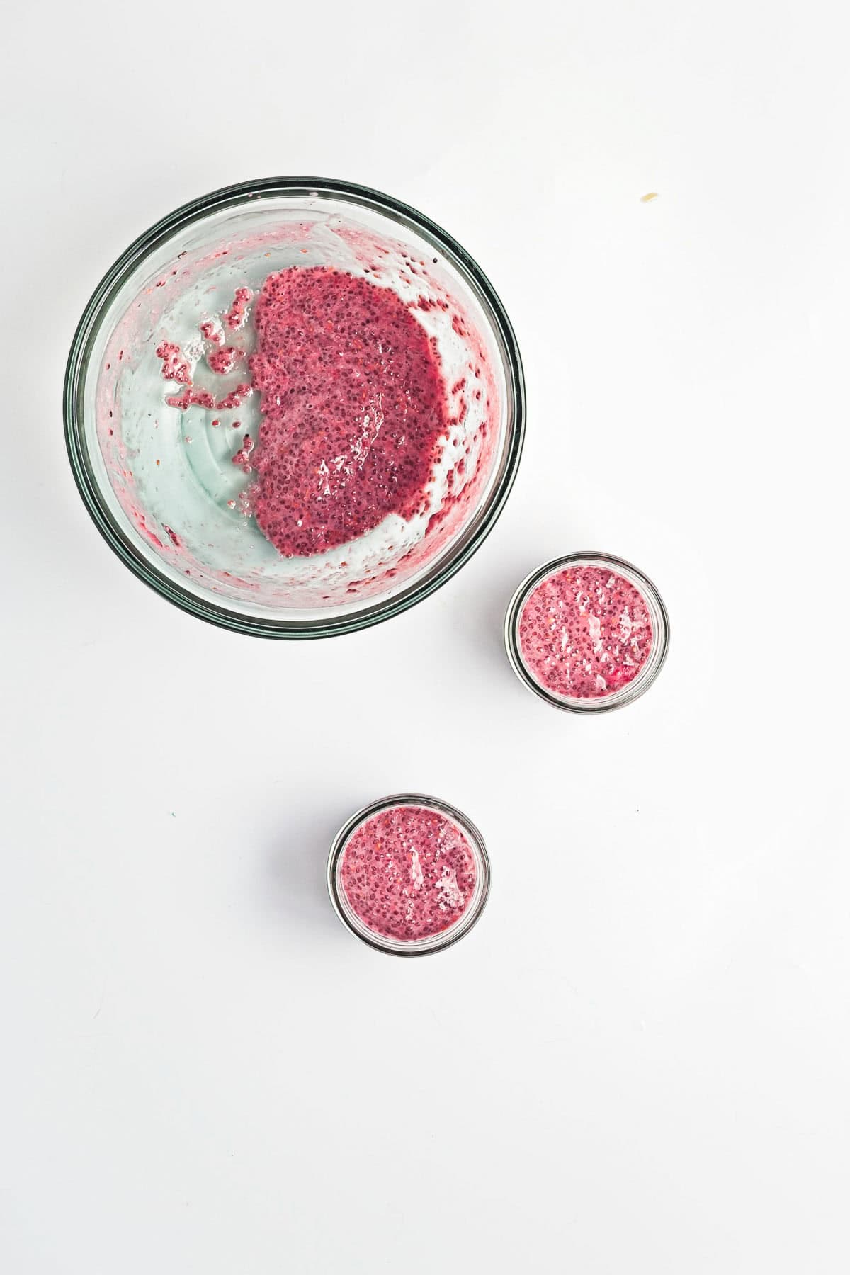 Overhead view of a large bowl and two small jars filled with Berry Chia Pudding, their pink hues vibrant against the white surface.