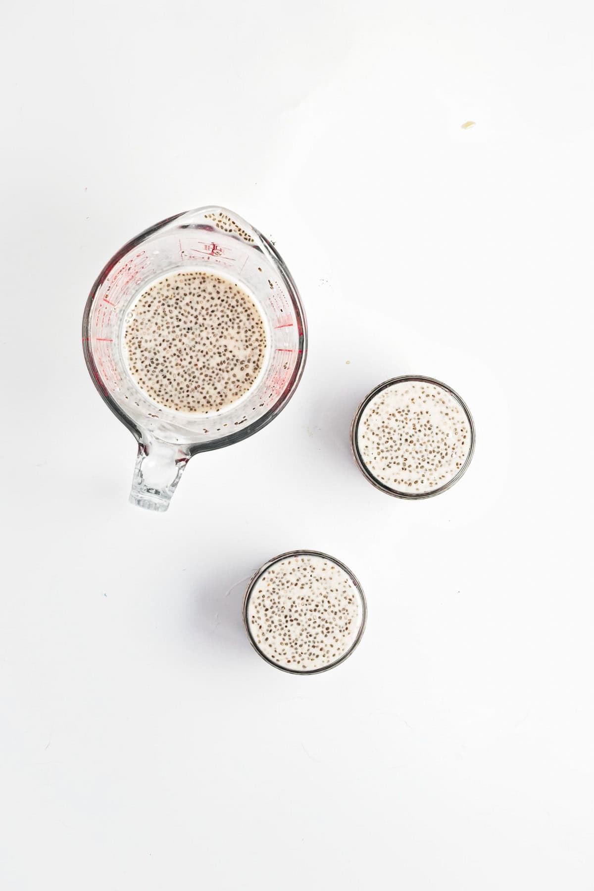 A measuring cup and two glasses filled with a creamy Berry Chia Pudding mixture on a white background.