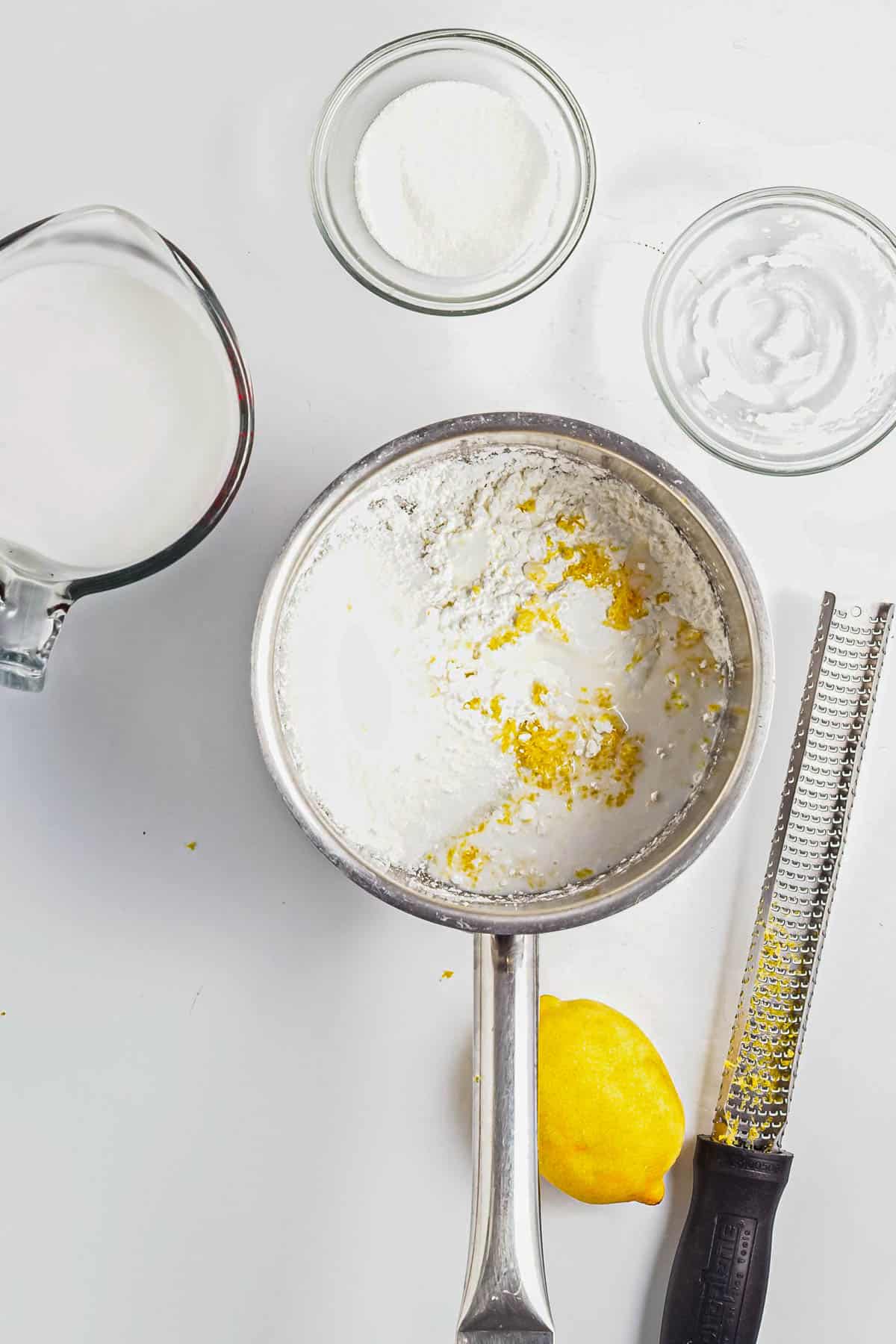 A saucepan with lemon zest, a lemon, grater, milk, and small bowls of sugar and cornstarch on a white surface-perfect for making Lemon Berry Coconut Custard.