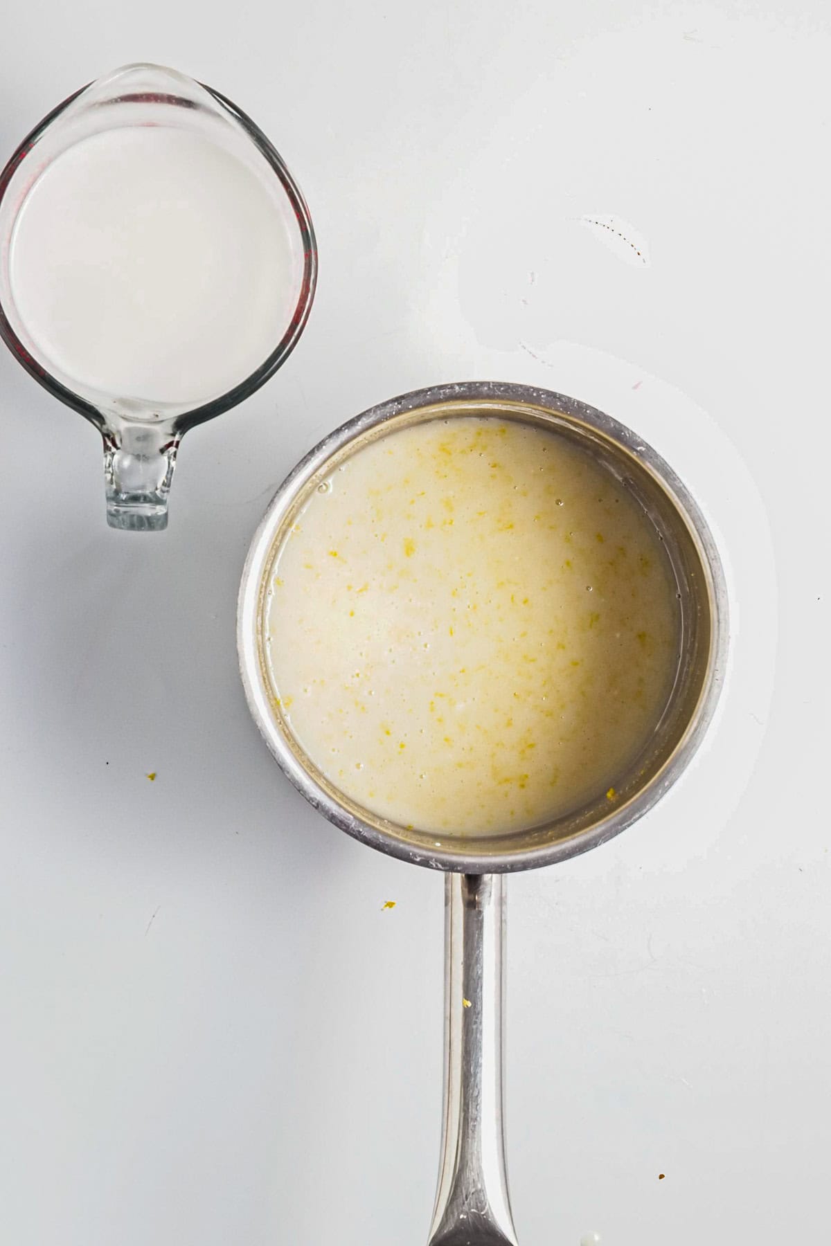A saucepan with creamy Lemon Berry Coconut Custard sits beside a glass measuring cup filled with milk on a white surface.