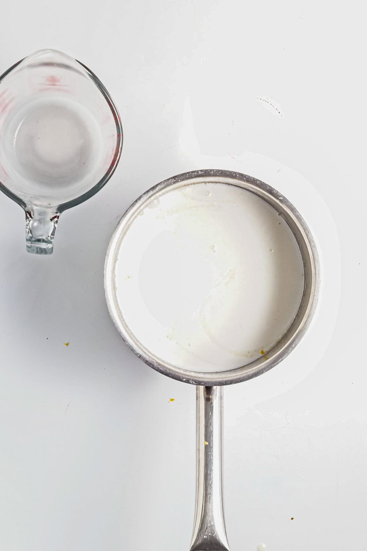 A saucepan with milk sits next to an empty glass measuring cup on a white surface, ready for preparing Lemon Berry Coconut Custard.