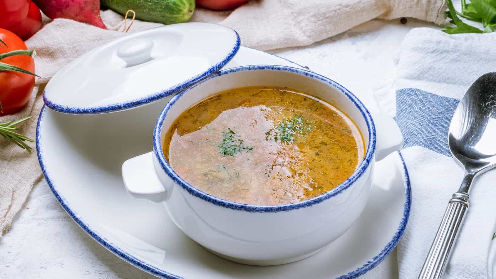 A white ceramic bowl with handles filled with brown soup, garnished with herbs, sits on a matching oval plate. A lid is partially open, and a spoon is placed nearby. Fresh vegetables are visible in the background.