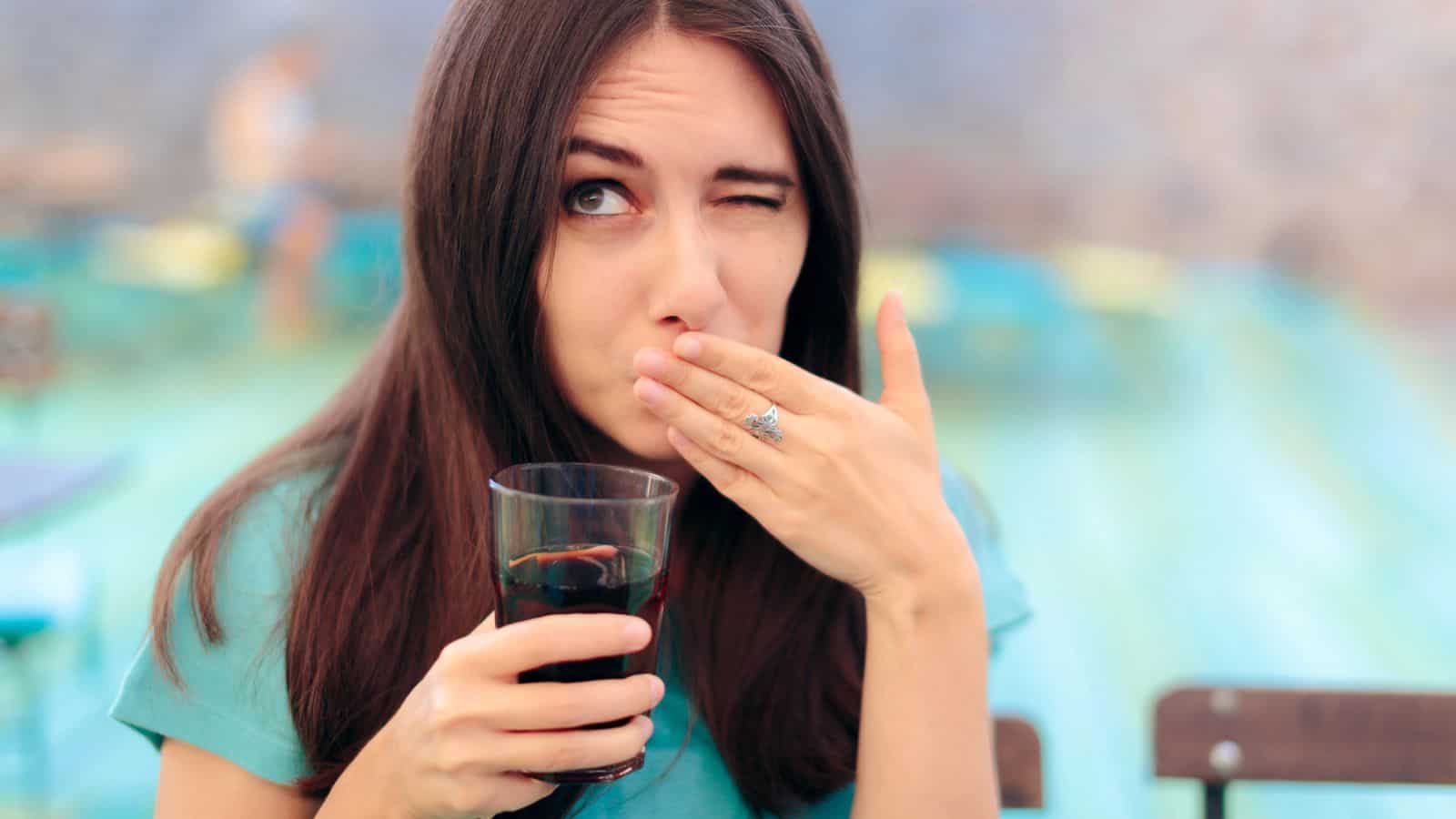 Woman holding a glass of dark drink, covering her mouth and winking outdoors.