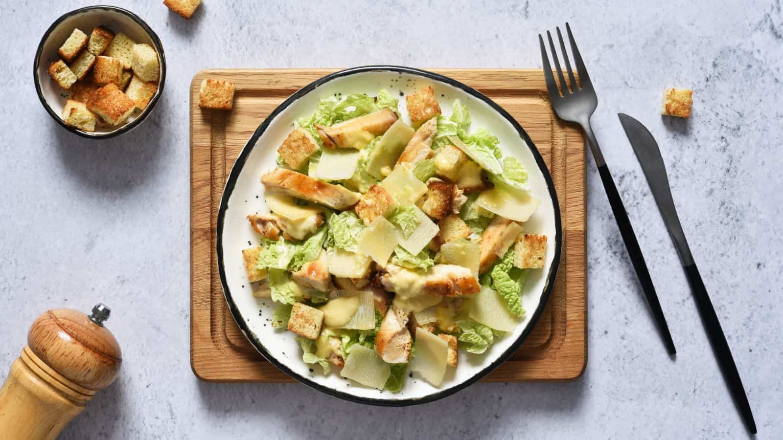A plate of Caesar salad with lettuce, grilled chicken, croutons, and parmesan shavings sits on a wooden board. A fork and knife are placed beside it, with a bowl of extra croutons and a pepper grinder nearby.