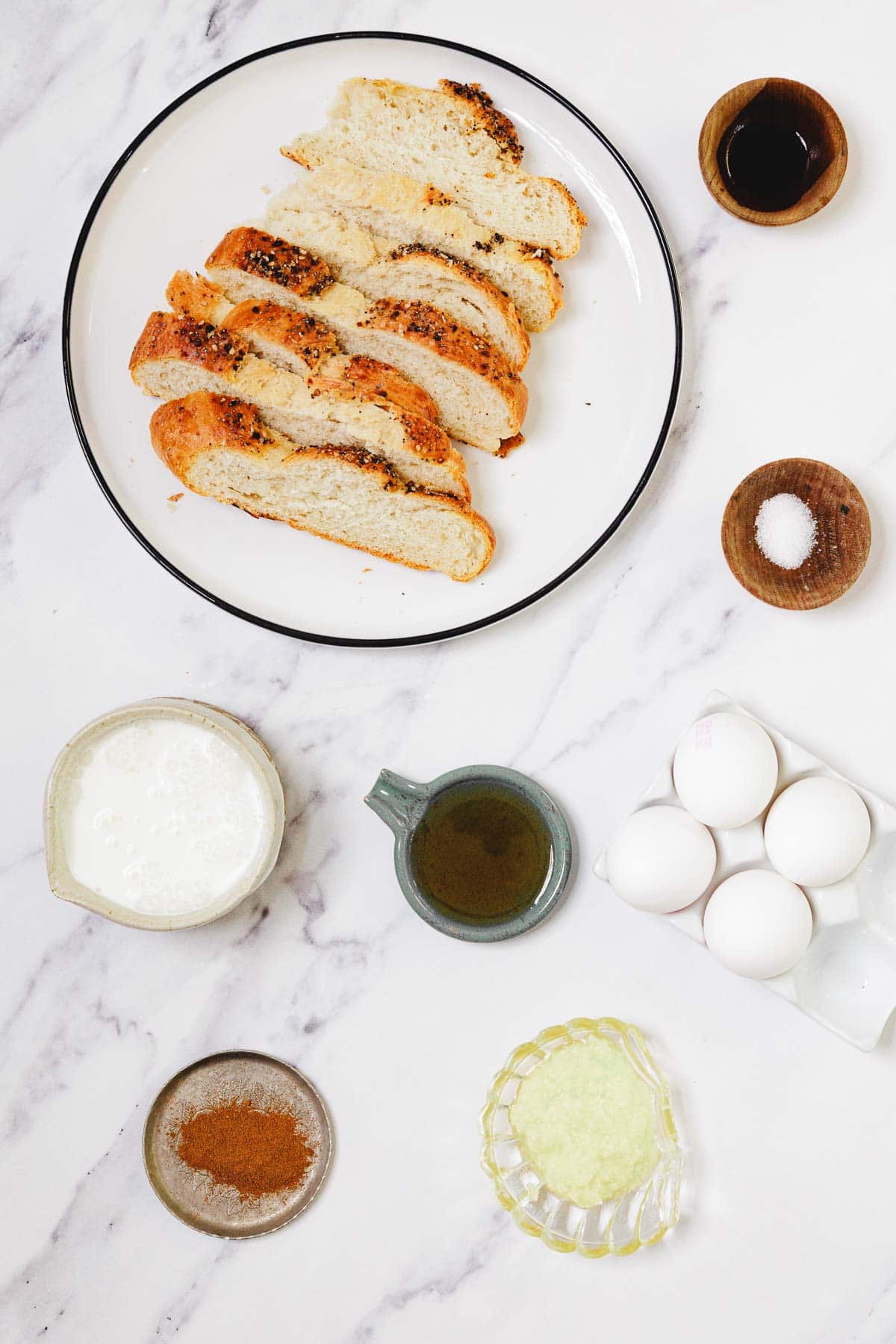 Sliced challah bread on a plate is surrounded by eggs, milk, oil, vanilla, cinnamon, sugar, and salt on a marble surface—perfect ingredients for delicious Challah French Toast.