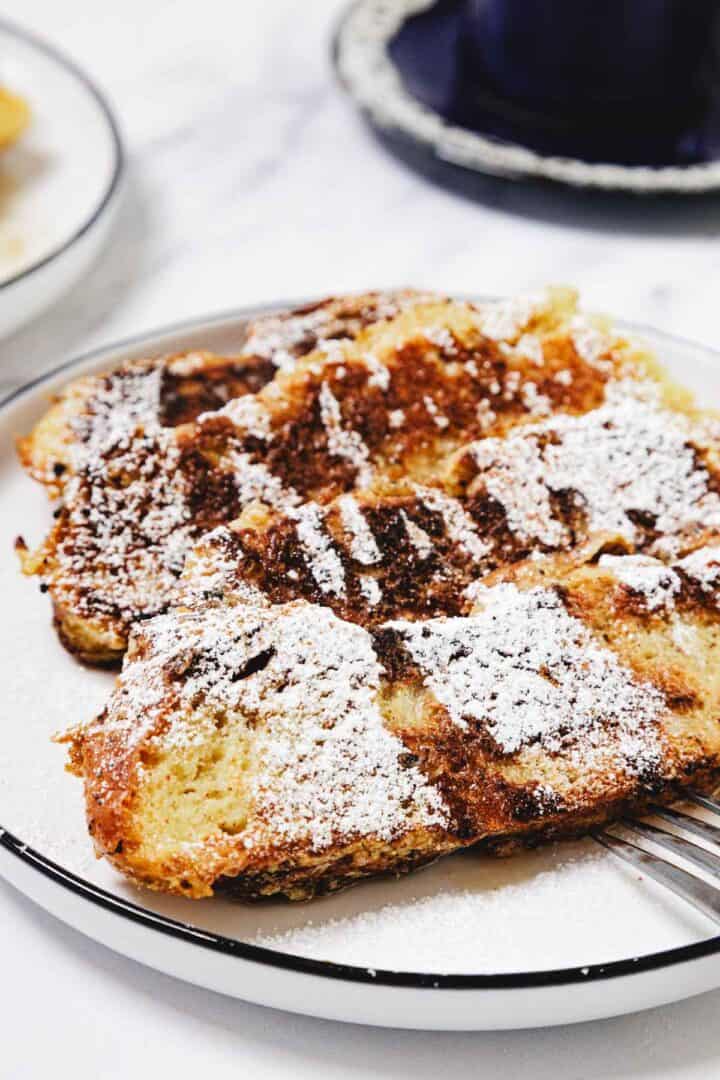 Two slices of Challah French Toast dusted with powdered sugar on a white plate with a fork.
